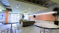 This single-floor space functions as a stylish communal lounge or lobby area, designed with a contemporary aesthetic that balances comfort and functionality. The layout is open and spacious, anchored by a seating arrangement of earth-toned sofas and lounge chairs that create an inviting social hub. Recessed ceiling lighting is complemented by a distinctive pendant light fixture with cylindrical glass elements, adding a refined focal point overhead.

A standout feature is the textured accent wall finished in warm terracotta tones, its wave-like relief pattern providing visual depth and artistic character to the space. A flat-screen television is mounted against this wall, enhancing the room's functionality as a gathering area. Adjacent high-top tables with modern acrylic seating offer an alternative setting for casual interactions.

Large windows and glass doors along one wall flood the interior with natural light, creating a bright and welcoming atmosphere while offering views to the exterior. The flooring transitions between tile and carpet, subtly defining different zones within the open plan. Overall, the space presents a polished, modern environment well-suited for social gatherings and relaxation.