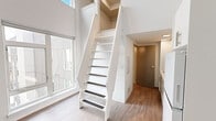 Located at 304 Puyallup Ave in Tacoma, WA, this thoughtfully designed studio makes exceptional use of its compact footprint through a smart loft-style layout. The main living area is immediately distinguished by soaring double-height ceilings that amplify the sense of space, while large windows flood the interior with abundant natural light and offer views of the surrounding neighborhood. Light wood-tone flooring flows throughout, complementing the clean white walls and creating a bright, airy ambiance.

A space-saving loft staircase with white-painted framing and dark-treaded steps connects the main floor to an upper loft area, providing valuable additional living or sleeping space. The efficient kitchen is outfitted with gray lower cabinetry, white upper cabinets, open shelving, a cooktop, microwave, and a full-size refrigerator with overhead storage — all designed to maximize functionality within the space.

The bathroom features wood-look tile flooring, a clean white vanity with sink, a toilet, and a shower/tub combination, all finished in a crisp, neutral palette. A dedicated entry door and thoughtful built-in storage throughout complete this well-appointed urban studio, ideal for those seeking modern, efficient living in the heart of Tacoma.