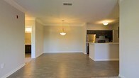 This single-floor space showcases an open-concept design that integrates the living and kitchen areas into one cohesive, functional layout. The living area is spacious and bright, featuring warm hardwood-style flooring that extends throughout the space, crisp white walls, and clean white baseboards that contribute to a fresh, modern aesthetic. A pendant light fixture hangs centrally, providing focused ambient lighting and serving as a subtle decorative element. The kitchen is positioned toward the rear of the open floor plan and is outfitted with dark espresso-toned cabinetry, a light-colored countertop peninsula that doubles as a breakfast bar or prep area, and stainless steel appliances including a refrigerator and stove. The dark cabinetry offers a striking contrast against the lighter tones of the surrounding space. A flush-mount ceiling light illuminates the kitchen zone. The overall ambiance is clean, contemporary, and move-in ready, with a neutral color palette that offers flexibility for a variety of interior styles. The open layout promotes a sense of connectivity and ease of movement throughout the main living areas.