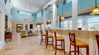 Located in Wesley Chapel, FL, the Point Cypress Blvd Clubhouse is a beautifully appointed single-story community gathering space designed with a coastal-inspired aesthetic. The interior is defined by dramatically tall ceilings with clerestory windows that flood the space with abundant natural light, accentuated by soft aqua-blue walls, white decorative columns, and elegant chandelier lighting.

The main gathering area features a prominent bar with a granite countertop and cushioned bar stools, seamlessly connected to a bright dining area furnished with round wooden tables and classic cross-back chairs in both white and cherry wood finishes. Tiled floors in warm travertine tones flow throughout, complementing the rich, layered décor.

Large windows dressed with decorative valances and teal drapery offer sweeping views of the outdoor pool and surrounding community, filling the space with a light, resort-like atmosphere. A comfortable lounge area features plush seating, a wall-mounted flat-screen television, shiplap-style accent walls, and curated coastal décor that creates an inviting, relaxed environment.

The clubhouse connects through glass-panel doors to adjacent areas and pool access points, making it a functional and stylish hub for community gatherings, socializing, and relaxation throughout the year.