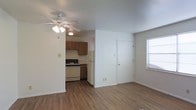 This single-floor unit offers a straightforward and functional layout designed for comfortable everyday living. The main living area is finished with warm hardwood-style flooring that extends throughout the space, lending a cohesive and clean aesthetic. A ceiling fan with an integrated light fixture is centrally mounted, providing both airflow and ambient lighting to the room. Natural light filters in through a window fitted with blinds, brightening the interior without sacrificing privacy. The entryway features a solid white front door, creating a clear and welcoming point of entry directly accessible from the living area. The kitchen sits adjacent to the living space in an open, semi-separated layout, making it easy to move between areas. It is equipped with warm-toned wood cabinetry offering ample storage, a gas range with oven, and a full-size refrigerator. The countertops and under-cabinet space provide a practical workspace for meal preparation. The neutral white walls throughout the unit create a versatile backdrop that suits a variety of interior styles. The overall ambiance is practical and unpretentious, offering a comfortable and move-in-ready living environment suited for those seeking a no-frills, functional space.