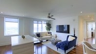 This well-appointed single-floor residence offers a thoughtfully designed open-concept layout that seamlessly blends functionality with modern elegance. The living area serves as the heart of the space, featuring rich hardwood flooring that adds warmth and sophistication throughout. Crisp white walls and a smooth ceiling create a clean, airy backdrop, while a ceiling fan ensures year-round comfort.

The living room is anchored by a large wall-mounted flat-screen television and complemented by a tufted cream sofa adorned with navy accent pillows, a sleek concrete-style coffee table, and a striking navy blue tufted accent chair — together creating a refined yet inviting atmosphere. A large window allows ample natural light to fill the room while offering open views of the surrounding landscape.

Adjacent to the living area, a kitchen counter with bar seating transitions smoothly into the open space, suggesting a connected dining and entertaining flow. Light wood cabinetry and a clean white countertop add a contemporary touch to this area. Lush indoor greenery provides a natural accent that softens the modern aesthetic. Overall, the space exudes a polished, urban sensibility with a warm and welcoming ambiance.
