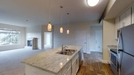 This well-appointed single-floor residence showcases a thoughtfully designed open-concept layout that seamlessly connects the kitchen, dining, and living areas. The kitchen serves as the heart of the home, centered around a generous island finished with elegant granite countertops and an undermount stainless steel sink. A trio of decorative pendant lights with warm amber tones hangs above the island, creating an inviting focal point while providing functional task lighting. The kitchen is further equipped with stainless steel appliances, including a dishwasher, and features white cabinetry with modern hardware throughout. Rich hardwood-style flooring anchors the main living areas, adding warmth and visual continuity to the space. The living area transitions to plush carpeting and is complemented by a classic fireplace with a white mantel, perfect for creating a cozy atmosphere. Neutral-toned walls throughout the residence provide a clean, versatile backdrop that suits a variety of interior styles. Interior doors are finished in crisp white, contributing to the bright and airy ambiance. The layout flows naturally between spaces, offering both functionality and comfort in a cohesive, move-in-ready setting.