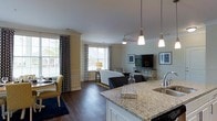This single-floor residence showcases a thoughtfully designed open-concept layout that seamlessly connects the kitchen and living areas, creating a spacious and inviting atmosphere. The kitchen is appointed with striking granite countertops, white cabinetry, a stainless steel sink, and pendant lighting that adds a contemporary touch overhead. The expansive island serves as both a functional workspace and a natural gathering point. Rich, dark hardwood flooring flows throughout the main living space, anchoring the design with warmth and sophistication. The living area is bright and airy, benefiting from multiple large windows dressed with patterned drapery that allow generous natural light to fill the room. A ceiling fan centered above the living space enhances comfort, while a media console provides a focal point for relaxation. Crown molding details along the walls and ceiling add a refined architectural element throughout. Decorative framed artwork and carefully chosen furnishings contribute to the polished, cohesive aesthetic. The overall ambiance is one of modern elegance balanced with everyday comfort, making this a welcoming and functional living environment.