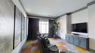 Located at 1971 Willow Loop Way in Knoxville, Tennessee, this single-floor amenity space serves as a dedicated media and lounge room designed for comfort and entertainment. The room is anchored by a row of plush, reclining theater-style chairs arranged to face a large flat-screen display mounted above a built-in media cabinet with gray cabinetry, offering ample storage and a clean, organized aesthetic. One of the most striking features of the space is the bold, multicolored modular carpet tiles underfoot, featuring a dynamic pattern of orange, gold, gray, and neutral tones that add energy and visual interest to the room. The walls are painted in a soft neutral tone and are accented with crown molding, giving the space a polished, finished look. A large window dressed with floor-length dark curtains allows natural light to filter in while maintaining an optimal viewing environment. A tall, lush indoor plant in the corner adds a refreshing natural element, softening the overall atmosphere. Wall-mounted artwork completes the décor, contributing to a curated, welcoming ambiance. The space is well-suited for group screenings, social gatherings, or quiet relaxation.