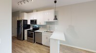 This single-floor space presents a cohesive, modern design characterized by light wood-look flooring and neutral grey-and-white tones carried consistently throughout. The kitchen serves as a standout feature, outfitted with a full suite of stainless steel appliances including a refrigerator, electric range, built-in microwave, and dishwasher. Crisp white cabinetry extends to the ceiling, providing ample storage, while a chevron-patterned tile backsplash adds a subtle decorative touch. White quartz countertops extend into a peninsula, offering additional workspace and casual seating potential. A modern pendant light and overhead track lighting illuminate the kitchen effectively. The bathroom maintains the same clean aesthetic, featuring a white vanity with quartz countertop, a toilet, and a glass-enclosed shower with a grab bar. An additional room, currently unfurnished, offers a versatile, clean-lined space with standard electrical outlets and a flush-mounted ceiling light. The 3D layout reveals a compact but well-organized floor plan with clearly defined zones for living, cooking, and bathing, all unified by consistent finishes and a bright, minimalist design sensibility.