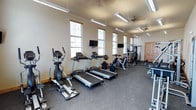 This single-level fitness center is designed to accommodate a comprehensive range of workout routines. The main cardio area lines one wall with multiple treadmills, an elliptical trainer, and stationary bikes, each positioned in front of wall-mounted televisions for an engaging workout experience. Natural light filters in through several windows fitted with blinds along both walls, contributing to an open and energizing atmosphere.

The strength training section is well-appointed with a cable machine, multi-function weight stations, and a full dumbbell rack offering an organized selection of free weights. Additional benches and functional training equipment are distributed throughout the space, providing versatility for varied exercise programs. A separate area near the entrance features vaulted ceilings and large windows that enhance the sense of openness, making it suitable for stretching or floor-based exercises.

Throughout the facility, durable rubber flooring provides both safety and practicality. Overhead lighting via hanging fluorescent fixtures, complemented by ceiling fans, ensures the space remains well-lit and comfortable during use. Rustic knotty pine doors add a warm architectural detail, and the neutral wall tones maintain a clean, focused environment conducive to fitness.