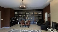 This single-floor space makes a strong visual impression through its thoughtful design and carefully curated details. Dominating the main wall is an expansive floor-to-ceiling built-in shelving unit finished in deep charcoal, providing ample display space for decorative objects, books, and accessories arranged throughout its multiple sections. The shelving creates a structured yet stylish backdrop that anchors the entire room.

At the center of the space, a round dining or meeting table is surrounded by patterned upholstered chairs featuring a rich red and black print, adding warmth and texture to the predominantly dark palette. Adjacent to the table, a dark armchair and sofa offer additional seating, reinforcing the room's functional versatility.

Overhead, a striking sculptural chandelier crafted from twisted branches and exposed bulbs serves as a standout focal point, complementing the space's bold character. Recessed lighting provides even ambient illumination throughout.

The light-toned tile flooring contrasts effectively with the darker furnishings, while a geometric patterned area rug in gray and white grounds the seating arrangement. A compact cabinetry unit along one wall adds practical storage, completing this well-appointed and design-forward interior.