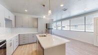 This single-floor space showcases a clean, contemporary design highlighted by light hardwood-style flooring that flows consistently throughout. The kitchen serves as a central focal point, featuring a large center island with a white quartz countertop, an undermount sink, and elegant pendant lighting overhead. Gray flat-front cabinetry lines the walls, complemented by a classic white subway tile backsplash and a full suite of stainless steel appliances, including a refrigerator, range, and built-in microwave. Recessed lighting adds to the bright, modern atmosphere.

The kitchen opens directly into a spacious living area bathed in natural light from a wide bank of windows fitted with horizontal blinds, offering views of the surrounding neighborhood. The bedrooms continue the light, airy aesthetic with the same flooring, recessed lighting, and generous windows.

Two bathrooms serve the space. One features a gray vanity with a white countertop, a large mirror, and a walk-in shower with sliding glass doors. The second bathroom includes a white vanity, a tub-shower combination, and matching flooring, both finished in a clean, neutral palette that reinforces the home's modern character throughout.
