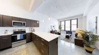 This thoughtfully designed single-floor space showcases an open-concept layout that seamlessly connects the kitchen and living areas, creating a fluid and functional environment. The kitchen is outfitted with rich dark-wood cabinetry, stainless steel appliances, and a generous island topped with a crisp white countertop — ideal for both meal preparation and casual dining. Pendant lighting hangs above the island, adding a warm, contemporary touch. The soaring ceilings lend an impressive sense of volume and openness throughout, while a track lighting system provides flexible illumination across the space. Polished concrete-style flooring runs throughout, contributing to the modern, urban aesthetic. The living area benefits from large, multi-pane windows that flood the space with natural light and offer outward views of the surrounding cityscape. The open floor plan allows for versatile furniture arrangements, making the living zone feel both spacious and inviting. The overall ambiance strikes a balance between industrial character — reflected in the exposed ceiling elements and raw finishes — and refined modern design, making this a compelling and highly livable space.