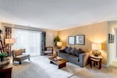 This single-floor residence offers a thoughtfully arranged layout with distinct living, dining, sleeping, and bathroom areas connected by an open corridor. The living room serves as the central gathering space, featuring warm golden-yellow accent walls, plush carpeting, and a large sliding glass door dressed with vertical blinds and decorative curtains that invite natural light and outdoor views. A generous sectional sofa, accent chairs, and a solid wood coffee table anchor the room comfortably.

The layout flows naturally into a separate dining area, illuminated by a ceiling fixture and furnished with a round table and upholstered chairs. The kitchen features rich dark-wood cabinetry, a double stainless steel sink, and laminate countertops providing ample workspace.

The two bedrooms differ notably in character. One presents neutral tones with carpet flooring, a large wall mirror over a dark wood dresser, and windows with patterned curtains. The second bedroom features a bold, dark accent wall, carpet flooring, and a window with views of surrounding greenery.

Two bathrooms complete the residence. One features warm yellow walls, a wood-vanity with white countertop, and a framed mirror. The second offers teal-painted walls, hardwood flooring, a full tub-shower combination with white tile surround, and a matching wood vanity.