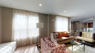 This single-floor space presents a thoughtfully designed interior that blends contemporary aesthetics with comfortable functionality. The living area is anchored by a tufted velvet sofa accented with vibrant orange and patterned throw pillows, complemented by a boldly upholstered armchair featuring an intricate floral motif in matching tones. Sheer floor-to-ceiling curtains dress multiple large windows, allowing generous natural light to filter throughout and casting warm, sunlit patterns across the light hardwood flooring. Recessed ceiling lighting and a sleek floor lamp provide additional illumination, while a glass-top coffee table and side tables add refined detail.

A mezzanine-style overlook features glass panel railings with stainless steel framing, offering views down to a lower common area accented by a stacked stone fireplace surround and decorative pendant lighting. The lower level incorporates a dedicated lounge zone with a wall-mounted flat-screen television set within a dark wood built-in media unit, supported by clean structural columns. Neutral gray wall tones and wide-plank hardwood flooring unify the space, creating a cohesive, modern ambiance throughout.