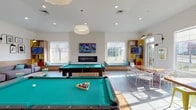 Located on Ashton Lane in Kent, Ohio, this single-floor community lounge space is designed with a modern, eclectic aesthetic that balances social activity with relaxed comfort. The open layout accommodates multiple distinct zones that flow seamlessly into one another, all unified by light hardwood-style flooring, crisp white walls, and abundant natural light streaming through large windows.

The game area serves as a central focal point, featuring two full-sized pool tables set beneath recessed lighting and a statement pendant fixture. A sleek linear fireplace is mounted on a textured accent wall, flanked by wide windows with built-in upholstered window bench seating dressed in teal cushions — ideal for casual lounging. Mounted flat-screen televisions are positioned throughout the space for viewing from multiple vantage points.

The lounge area offers generous sectional sofas in muted grey upholstery accented with colorful throw pillows, complemented by a curated gallery wall of framed pop-art-inspired prints and articulating wall-mounted sconce lighting. A dining nook features a live-edge wood table with contemporary white chairs, adjacent to a warm wood open shelving unit with vibrant yellow accents housing a television and decorative objects. The overall ambiance is welcoming, creative, and energetic.