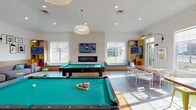 Located on Ashton Lane in Kent, Ohio, this single-floor community lounge space is designed with a modern, eclectic aesthetic that balances social activity with relaxed comfort. The open layout accommodates multiple distinct zones that flow seamlessly into one another, all unified by light hardwood-style flooring, crisp white walls, and abundant natural light streaming through large windows.

The game area serves as a central focal point, featuring two full-sized pool tables set beneath recessed lighting and a statement pendant fixture. A sleek linear fireplace is mounted on a textured accent wall, flanked by wide windows with built-in upholstered window bench seating dressed in teal cushions — ideal for casual lounging. Mounted flat-screen televisions are positioned throughout the space for viewing from multiple vantage points.

The lounge area offers generous sectional sofas in muted grey upholstery accented with colorful throw pillows, complemented by a curated gallery wall of framed pop-art-inspired prints and articulating wall-mounted sconce lighting. A dining nook features a live-edge wood table with contemporary white chairs, adjacent to a warm wood open shelving unit with vibrant yellow accents housing a television and decorative objects. The overall ambiance is welcoming, creative, and energetic.