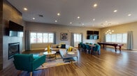 Located at 475 W Enterprise Dr in Mount Prospect, Illinois, this single-story community clubhouse is designed to offer a well-rounded gathering and recreation experience. The open-concept main area is anchored by a striking floor-to-ceiling tile-clad fireplace with a wall-mounted flat-screen television, creating a warm and inviting focal point. Rich hardwood-style flooring flows throughout, complementing the neutral walls and contemporary décor accented with bold teal and yellow furnishings. The lounge area features a generous sectional sofa, accent chairs, and a patterned area rug, all bathed in natural light from multiple windows dressed with patterned curtains and plantation shutters. Recessed lighting throughout ensures a bright, welcoming ambiance. A dedicated billiards area with a pool table and a modern globe chandelier offers additional entertainment, while a wall of mounted screens provides ample viewing. The full kitchen is equipped with stainless steel appliances, warm wood cabinetry, and a large wraparound breakfast bar with ample seating. A separate dining area with a long table and teal upholstered chairs accommodates group gatherings. A smaller private meeting room with French doors, a round table, and exterior views rounds out this versatile, amenity-rich space.