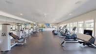 Located at 2401 Stanford Rd in Panama City, FL, this well-appointed fitness center offers a generously sized, single-floor layout designed to accommodate a comprehensive range of workout activities. The main exercise area is bright and open, featuring durable rubber flooring ideal for both cardio and strength training. Large windows line one wall, flooding the space with natural light and offering views of the surrounding outdoor area, including what appears to be a pool and palm-lined grounds — a distinctly Florida backdrop for workouts.

The facility is equipped with a robust selection of cardio machines, including treadmills, ellipticals, stationary bikes, and a rowing machine, all thoughtfully arranged to maximize floor space. A dedicated strength training zone features cable machines, weight benches, and various resistance equipment. An open stretch and functional fitness area is stocked with stability balls, yoga mats, and other accessories.

Full-length mirrors span an entire wall, enhancing the sense of space and allowing users to monitor their form. Mounted flat-screen TVs are positioned throughout for entertainment during workouts. Ceiling fans provide additional air circulation. The space also includes two closets, likely used for storage. The clean, white walls and neutral aesthetic create a motivating yet comfortable environment suited for all fitness levels.