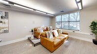 Located at 2986 W Lehman Ave in West Valley City, Utah, this single-floor community lounge offers a thoughtfully designed and efficiently utilized space. The room is finished with neutral-toned walls, plush carpet flooring, and clean white baseboards that contribute to a bright, modern aesthetic throughout.

The seating area is a central highlight, anchored by two tan leather sofas adorned with striped and solid accent pillows arranged to encourage conversation and relaxation. A wall-mounted flat-screen television serves as a focal point on one wall, complemented by an in-wall speaker system for an enhanced audio-visual experience. A small side table with a decorative plant adds a touch of greenery and warmth to this area.

Natural light filters in through large windows framed in dark metal, offering views of the surrounding outdoor landscape. Overhead, linear track lighting provides consistent, even illumination across the space.

A dining nook near the entrance features a round table with upholstered chairs and is accented by a set of framed abstract artworks on the adjacent wall. A glass-paneled door and window wall open visually into an adjacent fitness area, adding a sense of depth and transparency to the overall layout. The space is well-suited for casual gatherings, informal meetings, or quiet relaxation.