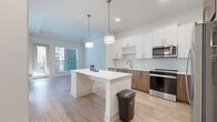 Located at 2124 American Industrial Way in Chamblee, GA, this well-designed one-bedroom, one-bathroom residence offers a clean, contemporary aesthetic across its single floor. The open-concept living and kitchen area serves as the heart of the unit, featuring light wood-style flooring that extends throughout the main living spaces. The kitchen is outfitted with stainless steel appliances, including a refrigerator, range, microwave, and dishwasher. A two-toned cabinetry design pairs white upper cabinets with warm wood-toned lower cabinets, complemented by a decorative scalloped tile backsplash and gold-accented hardware. A white center island with built-in shelving provides additional prep and storage space, while two pendant lights with brass fixtures hang above, adding a modern touch. A sliding glass door in the living area allows natural light to fill the space and provides access to an outdoor area. The bedroom is finished with plush wall-to-wall carpeting, neutral walls, and a window with blinds for natural light and privacy. A dedicated closet offers practical storage. The bathroom features a full tub-shower combo with tiled surround, a vanity, and consistent wood-style flooring, maintaining a cohesive look throughout the unit.