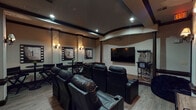 This private theater in Houston, TX is thoughtfully designed to deliver an authentic cinematic experience within a well-appointed, single-floor space. The room centers around a large wall-mounted flat-screen display positioned at the front, providing a clear sightline from every seat. Multiple rows of plush, dark leather power recliner chairs with built-in cup holders serve as the primary seating, offering comfort suited for extended viewing sessions. A dedicated bean bag seating area near the side walls provides a more casual alternative.

The interior design draws heavily from classic Hollywood aesthetics. Film-reel countdown artwork—numbered 1 through 4—lines the walls, framed in dark borders that complement the rich, warm color palette of tan and dark wood trim throughout. Decorative wall sconces cast a soft, ambient glow, enhancing the theater atmosphere while recessed ceiling lights provide functional overhead illumination. Dark structural columns and a curved ceiling detail add architectural depth to the space.

Along the side walls, director's chairs and bistro-style tables offer additional seating options. A classic popcorn machine stationed near the screen completes the moviegoing experience. Light hardwood flooring contrasts with the dark furnishings, contributing to the space's polished, inviting ambiance.