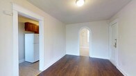 This single-floor space showcases a thoughtful blend of classic architectural details and functional design. The main living area features rich, dark hardwood flooring that adds warmth and character throughout. White walls and a flush ceiling light keep the space bright and neutral, providing a clean backdrop for furnishings.

A standout architectural feature is the rounded arched doorway, which adds a distinctive, classic touch to the interior and leads into an adjoining room that benefits from natural light through a visible window. This elegant detail gives the unit a sense of charm uncommon in standard floor plans.

To the left of the main living area, an opening leads directly into the kitchen, which is equipped with warm honey-toned wood cabinetry and includes a full-size white refrigerator. The transition between the kitchen and living area is open and accessible, promoting a connected feel between the two spaces.

Overall, the unit presents a well-maintained interior with a mix of timeless architectural elements—most notably the arched passageway—and practical amenities, making it a functional and characterful living space.