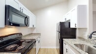 This single-floor residence presents a bright, neutral interior designed for comfortable everyday living. The open living area is a standout feature, offering plush carpeting, a classic fireplace set against tiled surround, and large sliding glass doors with vertical blinds that invite natural light and provide access to the outdoors. A peninsula with a granite countertop and pendant lighting connects the living space to the kitchen, creating a functional and visually cohesive layout.

The galley-style kitchen is well-appointed with crisp white shaker cabinetry, matte black hardware, granite countertops, and light wood-tone flooring. Black appliances — including a refrigerator, electric range, and over-the-range microwave — complement the clean, modern aesthetic. Ample upper and lower cabinet storage lines both sides of the kitchen.

The bedrooms are carpeted and finished in soft, neutral tones, each offering natural light through windows with blinds. One bedroom features a built-in ledge detail beneath the window, adding a subtle architectural touch.

The bathroom is finished with white subway tile in the tub/shower surround, matte black fixtures, wood-look tile flooring, and white cabinetry — maintaining a cohesive, contemporary feel consistent with the rest of the home.