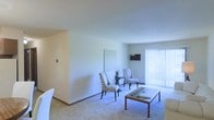 This single-floor space offers a comfortable and functional layout with a focus on simplicity and practicality. The living area serves as the centerpiece of the space, featuring soft wall-to-wall carpeting that adds warmth underfoot and a neutral color palette across the walls and ceiling that creates a calm, inviting atmosphere. Natural light filters generously into the room through a large window dressed with vertical blinds, allowing for flexible light control throughout the day. A wall-mounted climate control unit ensures year-round comfort. The open layout of the living area provides ample room for a variety of furniture arrangements, as evidenced by the patterned accent chairs and a sleek coffee table that bring a touch of personality to the otherwise understated interior. Clean baseboards and smooth walls throughout reflect a well-maintained environment. The overall ambiance is bright, airy, and uncluttered, making the space feel open and easy to personalize. This is a straightforward, move-in-ready space ideal for those who value a clean aesthetic and practical living on a single, accessible level.