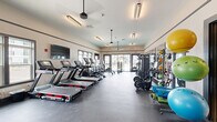 Located at 100 Dorchester Dr in Lumberton, NJ, this well-equipped fitness center offers a comprehensive workout environment within a single, open-plan floor. The space is designed with functionality and comfort in mind, featuring durable rubber flooring throughout that provides both traction and impact absorption during exercise.

The cardio zone is prominently lined with a row of modern treadmills and elliptical trainers, each equipped with individual digital displays. Complementing these are rowing machines and stationary cycling bikes, offering a diverse range of aerobic options. A strength training area includes a multi-functional cable machine, adjustable benches, and a full dumbbell rack, catering to a variety of resistance training needs.

Large windows along multiple walls flood the interior with natural light while offering views of the surrounding community. Floor-to-ceiling mirrors expand the visual space and support proper form during workouts. A dedicated accessory station stocks yoga mats, resistance bands, and stability balls for functional and flexibility training.

Overhead, pendant-style ceiling fans ensure consistent airflow throughout the space. Wall-mounted flat-screen televisions provide entertainment during cardio sessions. The overall aesthetic is clean and contemporary, with neutral walls accented by dark trim detailing, creating an energizing yet focused atmosphere ideal for daily fitness routines.