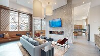 This thoughtfully designed single-floor communal space offers a generous, open-plan layout that seamlessly blends a lounge, dining, and kitchen area into one cohesive environment. The soaring ceilings lend an airy, expansive quality to the interior, accentuated by oversized cylindrical pendant lights and geometric diamond-frame light fixtures that serve as striking focal points overhead.

At the heart of the lounge, a dramatic floor-to-ceiling textured tile accent wall frames a sleek linear fireplace and a wall-mounted display screen, creating an inviting gathering area furnished with plush blue sofas, leather sling chairs, and a layered area rug. Flanking this zone, a built-in wood shelving unit with triangular geometric detailing adds warmth and visual interest.

The dining area features warm wood tables paired with curved orange upholstered chairs and built-in window bench seating. Tall, angled wood-framed windows flood the space with natural light while providing an architectural design element that defines the room's character.

The kitchen is outfitted with rich wood-grain cabinetry, a vibrant teal mosaic tile backsplash, crisp white countertops, a waterfall-edge island with bar seating, and a stainless steel range hood. Gold-toned ring pendant lights add a refined finishing touch. Large-format slate-toned tile flooring unifies the entire space, contributing to its modern, sophisticated ambiance.