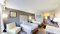 Located at 1400 W Tunnel Blvd in Houma, Louisiana, this hotel room offers a practical and comfortable layout designed to accommodate guests with ease. The main sleeping area features two queen-sized beds dressed in crisp white linens, each backed by large upholstered headboards framed in warm wood tones. A shared nightstand positioned between the beds holds a lamp and telephone, while a cushioned armchair near the window provides a relaxing spot to unwind. Natural light filters in through a window dressed with layered curtains, giving the room a bright and welcoming feel. The walls feature a two-tone design with a textured gray accent wall complementing the lighter upper portions, creating a clean, contemporary aesthetic. Hardwood-style flooring runs throughout, adding warmth to the space. On the opposite end of the room, a wooden dresser supports a flat-screen television, accompanied by a compact mini-refrigerator and microwave below, along with a dedicated work desk and chair — ideal for business travelers. The bathroom is efficiently designed with a granite-topped vanity, a large framed mirror with overhead lighting, and a separate wet area housing a combined tub and shower with tile surround, toilet, and towel storage.