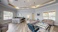 This single-floor gathering space is thoughtfully designed to blend sophistication with comfort. The main lounge area is anchored by a striking tray ceiling with integrated ambient lighting and a ceiling fan, adding both architectural interest and a warm glow to the room. Light grey walls, crown molding, and light wood-toned flooring run throughout, creating a cohesive and inviting atmosphere.

The seating area is generously proportioned, accommodating multiple sofas, accent chairs, and a glass coffee table atop a patterned area rug. An arched interior doorway adds a classic architectural detail, while large windows fitted with plantation shutters invite natural light and offer views of the outdoor pool area.

A wet bar with rich wood cabinetry, dark granite countertops, an undermount sink, and bar seating provides a functional and stylish amenity for entertaining. A decorative mosaic mirror above the bar counter serves as a notable design accent.

Opposite the lounge, a wall-mounted flat-screen television is paired with a media console, offering a comfortable entertainment zone. French doors lead directly to the outdoor pool area, seamlessly connecting the interior with the exterior. Additional louvered bi-fold doors provide access to utility or storage areas. Recessed lighting throughout the space ensures consistent illumination across all zones.