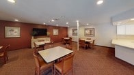 Located at 400 Luella St N in Saint Paul, Minnesota, this single-floor community space is thoughtfully designed to serve as a shared gathering hub. The centerpiece is a spacious dining and activity room, finished with warm patterned carpeting and accented by deep red and neutral-toned walls. Multiple dining tables with cushioned chairs are arranged throughout, providing ample seating for group meals or social events. Recessed lighting illuminates the room evenly, while framed artwork and a wall-mounted flat-screen television add a welcoming, residential character. An adjacent kitchen is outfitted with white cabinetry, light countertops, and white appliances including a range with overhead microwave and a dishwasher, all complemented by light hardwood-style flooring. A connected common area features patterned carpeting, large windows that invite natural light, and access to glass-paneled entry doors, creating an open and inviting transition space. A water cooler station and seating nook are also present. Additional rooms provide supplemental gathering or meeting space with windows fitted with vertical blinds and individual climate control units. Overall, the space offers a functional, comfortable, and community-oriented environment well-suited for shared living or residential programming.