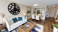 This single-floor residence offers a thoughtfully designed open-concept layout that seamlessly connects the living, dining, and kitchen areas. The living room is bright and inviting, featuring rich hardwood-style flooring, a large sliding glass door that floods the space with natural light, and access to an outdoor area. A bold gear-style wall clock serves as a striking focal point, complementing the contemporary furnishings and patterned area rug.

The kitchen is well-equipped with stainless steel appliances, including a refrigerator, microwave, and range. Two-tone cabinetry — white uppers and grey lowers — is paired with crisp white quartz countertops and a classic subway tile backsplash, creating a clean, modern aesthetic. A breakfast bar connects the kitchen to the dining area, which comfortably accommodates a full dining set.

The bedroom is a serene retreat with plush carpet flooring, a window providing natural light, and a wall-mounted air conditioning unit. Ample storage is provided by a dedicated walk-in closet featuring wire shelving throughout.

The bathroom is finished in a clean, neutral palette with a full tub-shower combination and brushed nickel fixtures. Throughout the space, neutral wall tones and cohesive design choices create a warm and functional living environment.