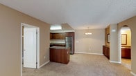 This upstairs unit at 1617 Live Oak in Vista offers a well-organized, single-floor layout that connects living, dining, and kitchen areas in an open, functional arrangement. The kitchen is a standout feature, outfitted with rich dark-wood cabinetry, granite-style countertops, a stainless steel refrigerator, a stainless steel range with microwave above, and a built-in peninsula that provides additional prep space and defines the transition into the living area. Open corner shelving adds a practical design detail. A multi-bulb chandelier pendant light illuminates the adjacent dining area, which flows seamlessly into the spacious carpeted living room. The living room offers generous floor space and includes a sliding glass door with vertical blinds, providing access to natural light. The bedroom features plush carpeting and mirrored sliding closet doors, maximizing the sense of space. A second room with carpeting and a wall-mounted thermostat offers additional flexibility. The bathroom is appointed with dark wood vanity cabinetry, granite-style countertops, a full tub-shower combination, and tile flooring. Textured ceilings and warm neutral-toned walls are consistent throughout the unit.