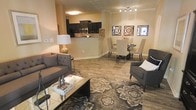 This single-floor residence showcases an open-concept design that seamlessly integrates the living, dining, and kitchen areas into one cohesive and functional space. The living area is anchored by warm wood-look flooring and features tufted upholstered seating, a glass-top coffee table, and a patterned area rug that adds texture and visual interest underfoot. Track lighting overhead provides even illumination across the space.

The dining area transitions naturally from the living room, accommodating a round table with upholstered chairs and decorative wall art that adds a curated touch to the neutral-toned walls. Adjacent to the dining space, the kitchen is defined by dark cabinetry contrasted against a raised breakfast bar with light-toned countertops, creating a natural boundary without sacrificing the open feel. Stainless steel appliances, including a microwave and range, are visible within the kitchen, contributing a modern finish.

The warm beige walls and consistent flooring unify the entire space, creating a cohesive and inviting atmosphere throughout this well-designed, move-in-ready layout.