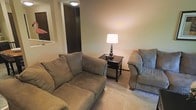 This single-floor space centers around a comfortable and understated living area designed for relaxed everyday living. The sitting area is furnished with a plush, neutral-toned sofa and a matching upholstered armchair, both finished in a soft gray-green fabric that complements the warm beige tones of the walls and carpet. A dark-finished end table positioned between the seating provides a functional surface, topped with a decorative globe-based lamp that casts a warm, ambient glow throughout the room. The carpeted flooring adds a sense of softness and warmth underfoot, contributing to the overall cozy character of the space. The walls are smooth and painted in a light, neutral tone, creating a calm and uncluttered backdrop. A flush-mount ceiling light provides additional overhead illumination. Adjacent to the sitting area, a dark-paneled door or closet element adds a subtle contrast to the lighter surroundings. The layout is practical and efficiently arranged, making good use of the available floor space. Overall, the space presents a modest, functional, and welcoming environment well-suited for quiet relaxation or casual everyday use.