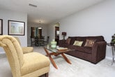 This single-floor residence offers a thoughtfully designed layout that balances comfort and functionality throughout. The living area provides a welcoming atmosphere with soft carpeting, neutral walls, and window treatments that allow for natural light, creating a relaxed and inviting ambiance. Adjacent to the living space, a dedicated dining area accommodates a round table set beneath a pendant light fixture, complemented by decorative wall art and an open passage to the kitchen. The kitchen is outfitted with rich wood cabinetry, a gas range with a ventilation hood, tile backsplash, and ample counter space, making it both practical and visually cohesive. The primary bedroom is generously sized with plush carpeting, a large window with outdoor views, and direct access to an en-suite bathroom featuring a wide vanity with a single sink, wood cabinetry, and a full mirror. A second bedroom also offers comfortable proportions, carpeted flooring, and natural light. The second bathroom mirrors the warm aesthetic of the primary bath, complete with wood-toned cabinetry, a vanity sink, and a shower-tub combination. Throughout the residence, a consistent palette of neutral tones and warm wood accents ties each space together cohesively.