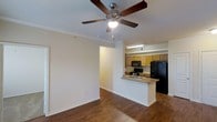 This single-floor residence offers an open and cohesive layout that seamlessly connects the living area to the kitchen. The living space features rich hardwood flooring that extends throughout, contributing warmth and visual continuity across the room. Crown molding lines the ceiling, adding a refined architectural detail, while a five-blade ceiling fan with an integrated light fixture serves as both a functional and decorative centerpiece overhead.

The kitchen is positioned at the rear of the open-plan space and is defined by a peninsula with a built-in sink, providing a natural boundary between the cooking and living areas while also offering additional counter space. Medium-toned wood cabinetry lines the upper and lower kitchen walls, providing ample storage. Black appliances, including a refrigerator, microwave, and range, provide a consistent and practical finish throughout the kitchen. Recessed fluorescent lighting above the kitchen ensures the workspace is well-lit.

The neutral, light-toned walls throughout the space create a clean and versatile backdrop, lending an airy, open ambiance well-suited to a variety of interior styles.