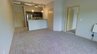 This single-floor space showcases an open-concept design that seamlessly connects the living area and kitchen, creating a bright and functional layout. The living space is finished with plush wall-to-wall carpeting in a neutral tone, lending warmth and comfort underfoot. A peninsula-style kitchen counter divides the spaces while maintaining an open sightline throughout. The kitchen is well-appointed with stainless steel appliances, including a refrigerator, microwave, and range, complemented by dark cabinetry and granite-style countertops. Industrial-style exposed ductwork runs along the ceiling, adding a contemporary urban character to the space. Pendant lighting hangs above the kitchen counter, providing focused task lighting with a modern aesthetic. The walls are finished in a soft warm tone, contributing to an inviting ambiance. Multiple interior doors lead to additional rooms, suggesting private spaces such as bedrooms or a bathroom, offering separation and versatility. The overall design balances open communal living with practical, private areas, making it a well-rounded and thoughtfully arranged space suited for everyday living.