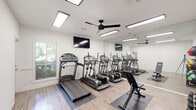 Located at 3214 Brassfield Rd in Greensboro, NC, this fitness center offers a well-appointed, single-room workout space designed to maximize functionality. The room features light wood-look flooring throughout, crisp white walls, and a clean, modern aesthetic that creates an open and energizing atmosphere.

The cardio zone is equipped with multiple treadmills and elliptical machines arranged in a row, complemented by wall-mounted flat-screen TVs for entertainment during workouts. A full-length wall mirror spans one side of the room, visually expanding the space and assisting with form during exercise. A ceiling fan keeps the air circulating overhead, while recessed and surface-mounted LED panel lighting ensures the room remains bright and well-lit at all times.

The strength and accessory area includes a dumbbell rack with a full set of free weights, a medicine ball wall rack with color-coded balls, and a adjustable workout bench, providing options for a variety of strength training exercises. A sanitation station with wipe dispensers is conveniently located near the entrance, alongside a posted Fitness Center Rules placard. A window along one wall allows natural light in and offers a view of the surrounding greenery. The space is practical, clean, and thoughtfully organized for an efficient fitness experience.
