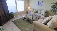 This single-floor space features a inviting living room designed with a cohesive, neutral color palette. The room is anchored by plush, beige upholstered seating, including a sofa adorned with decorative green and cream accent pillows, complemented by a matching armchair. A glass-top coffee table with elegant curved wooden legs serves as a central focal point, accented by tall candlestick holders and set atop a rich, olive-green area rug that adds warmth and texture to the carpeted floor. Natural light flows generously into the room through large windows dressed with white horizontal blinds, brightening the soft sage-green walls. A floor lamp with a classic cream shade provides additional ambient lighting, while a framed botanical print on the wall adds a decorative touch. The overall ambiance is calm and welcoming, with a tasteful blend of natural tones and carefully chosen furnishings that create a comfortable and functional living environment.