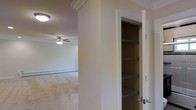 This single-floor space presents a clean, functional layout highlighted by neutral tones throughout. The living area features wall-to-wall carpet and is equipped with a ceiling fan with a light fixture, providing both comfort and illumination. Crown molding along the upper walls adds a refined architectural detail to the otherwise understated interior. A built-in closet, visible through an open white-framed door, offers multiple wire shelving tiers, providing well-organized vertical storage space. The walls are painted in a soft, warm neutral tone that contributes to a calm and inviting ambiance. Baseboard heating is present along the perimeter walls, indicating a practical heating solution integrated seamlessly into the design. The open passageway between areas allows for a natural flow through the space, enhancing the sense of connectivity within the floor plan. Overall, the space is modest yet functional, combining practical storage solutions with comfortable living features in a straightforward, well-maintained interior.