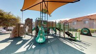 This outdoor recreational space is designed to serve a wide range of ages and activity levels, combining a feature-rich children's playground with a dedicated open-air fitness area. The centerpiece of the playground is a multi-level structure with a nature-inspired aesthetic, incorporating tree trunk-style climbing elements, log-themed panels, and earthy tones of brown and green. Children can enjoy multiple slides, including a twin-lane slide and a standalone wavy slide, along with a rock climbing wall fitted with colorful handholds. A separate boulder-style climbing structure adds additional challenge. Generous tan shade sails are suspended overhead, providing meaningful sun protection across the play area. The entire playground sits on a cushioned rubberized surface in a soft teal hue, prioritizing safety. Adjacent to the playground is a well-appointed outdoor fitness zone equipped with a variety of green and brown exercise stations, including parallel bars, balance beams, and stretching equipment, all set on the same rubberized flooring. The area is enclosed by chain-link fencing and surrounded by mature trees and palm trees, lending a shaded, park-like atmosphere. Residential buildings with terracotta rooflines form the backdrop, situating this recreational space within a community setting.