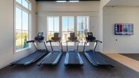 Located at 3030 Barrow Rd. in Charlotte, NC, this single-floor fitness center is designed to support a comprehensive workout experience in a bright, well-appointed environment. The facility features soaring ceilings with exposed beam detailing, flooding the space with an open, airy atmosphere accentuated by expansive floor-to-ceiling windows that invite abundant natural light and outdoor views of the surrounding landscape.

The cardio zone is equipped with multiple treadmills positioned directly in front of large windows, providing natural scenery during workouts. Additional cardio equipment, including a stair climber and recumbent bike, is arranged in a separate section beneath a dramatic wall of windows framed by bold architectural beams. The strength training area offers a cable machine system, a Smith machine with a bench press, and a well-stocked dumbbell rack displayed alongside a round wall mirror, catering to both free-weight and machine-based training.

Durable rubber flooring covers the workout areas, transitioning to a polished aggregate floor in the lounge zone. This relaxation area features a wall-mounted television, a built-in refreshment counter with a beverage refrigerator and wine rack, and a glass-paned door connecting to the main fitness floor. The clean, neutral color palette of white walls and dark trim gives the space a polished, contemporary aesthetic throughout.