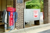 This single-level outdoor amenity area serves as a practical, dual-purpose facility within a residential community. The covered structure is supported by brick columns and features a clean white ceiling outfitted with fluorescent lighting, ensuring visibility during all hours of use. The concrete floor includes an integrated floor drain, making cleanup efficient after use.

A standout feature of this space is the stainless steel pet washing station, equipped with a built-in step for easy pet access and a mounted hose for convenient rinsing. Adjacent to this is a dedicated car wash bay — a generously sized, open-air covered area with posted instructional signage on the brick half-wall. The open sides of the structure provide unobstructed views of the surrounding mature tree line, lending a natural, serene backdrop to the utilitarian space.

The facility borders a spacious surface parking lot with traditional lamp posts and landscaped islands. The broader community setting includes multi-story residential buildings visible in the distance. This amenity space offers residents convenient, well-equipped options for vehicle and pet care within easy reach of their homes.