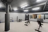 Located at 1 Websters Landing in Syracuse, NY, this exercise room offers a well-equipped fitness facility designed to support a comprehensive workout experience. The space is divided into distinct functional zones, making efficient use of its generous floor area across a single level.

The cardio zone features multiple treadmills, elliptical machines, and a stationary bike, all lined along a mirrored wall that visually expands the room and allows users to monitor their form. A rack of dumbbells in graduated weights is conveniently positioned nearby for quick transitions between exercises.

The strength and free-weight area includes a multi-function cable machine, an adjustable workout bench, and a leg curl/extension station, providing options for both compound and isolated strength training movements. Additional accessories such as resistance bands and stability balls are also available.

Architecturally, the space features an exposed industrial-style ceiling with visible ductwork and steel beams painted in dark tones, contrasted by bright fluorescent strip lighting that ensures thorough illumination throughout. Structural columns add to the industrial character of the room, while a section of original exposed brick and stone wall introduces a striking textural element. The floor is finished with durable, light-toned rubber flooring suited for high-traffic fitness use. The overall ambiance is modern, functional, and utilitarian, befitting a well-maintained fitness facility.