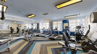 Located at 7029 W Friendly Ave in Greensboro, North Carolina, this fitness center offers a thoughtfully designed exercise space equipped for a comprehensive workout experience. The room is outfitted with an impressive array of cardio equipment, including multiple treadmills, elliptical trainers, stationary bikes, and recumbent cycles, all arranged for easy access and efficient use of the floor space. Strength training needs are addressed with a multi-station cable machine, weight benches, and free weights. The interior design strikes a bold visual balance, featuring deep navy blue accent walls contrasted against light gray and white walls adorned with crisp crown molding. A vibrant geometric-patterned carpet in tones of teal, gold, gray, and brown anchors the space and adds a contemporary energy to the room. Recessed rectangular light fixtures with warm yellow panels illuminate the ceiling, complementing the abundant natural light that streams in through multiple large windows along the perimeter walls. Full-length mirrors along one wall visually expand the space and allow exercisers to monitor their form. A wall-mounted clock and posted fitness center rules reflect the room's practical, user-focused design. The overall ambiance is modern, energetic, and welcoming.