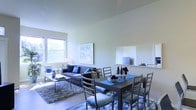 This single-floor space offers a thoughtfully designed open-concept layout that combines the living and dining areas into one cohesive, inviting environment. The living area is anchored by a plush, dark-toned sofa accented with blue cushions, set atop a layered area rug that adds texture and warmth to the light hardwood-style flooring. A sleek coffee table sits at the center, complementing the understated, modern aesthetic throughout. Large windows allow abundant natural light to flood the space, enhancing the airy feel of the neutral-toned walls and high ceilings. A framed art piece adds a subtle decorative touch to the living zone. The dining area features a sturdy dark wood table surrounded by cushioned ladder-back chairs, providing comfortable seating for gatherings. A built-in shelving niche along the wall offers a dedicated space for a wall-mounted display and decorative accents, adding architectural interest to the room. The overall ambiance is clean, contemporary, and well-balanced, with a harmonious blend of cool blue tones and warm neutrals that create a welcoming and functional living environment throughout.