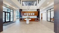 This contemporary single-floor space is designed with a strong emphasis on modern architecture and refined finishes. The grand entrance lobby immediately commands attention with its soaring double-height ceiling, expansive skylights, and a striking geometric chandelier that serves as a central focal point. Large-format neutral stone tile flooring spans the open reception area, anchored by a sculptural white pedestal table. A full-length walnut millwork wall with illuminated glass backsplash and curated artwork provides an elegant backdrop.

Black-framed glass partitions divide the leasing office area, maintaining visual openness while defining functional workspaces. A well-appointed lounge area features plush seating, wood slat accent walls, wall-mounted displays, and a sliding barn door, creating a warm yet contemporary atmosphere. Floor-to-ceiling windows flood this area with natural light and offer panoramic outdoor views.

The kitchen and social bar area is outfitted with a generously sized walnut island with white countertops, backlit glass tile, a wood slat ceiling detail, pendant lighting, and a row of modern bar stools. Open shelving and built-in cabinetry provide functional storage throughout. Together, these areas create a cohesive, upscale environment suited for both professional and social interaction.