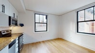 This single-floor unit showcases a clean, contemporary design characterized by crisp white walls, light hardwood flooring, and bold black-framed windows that frame urban cityscape views throughout. The open-plan kitchen is a standout feature, outfitted with sleek white flat-front cabinetry, granite countertops, a classic white subway tile backsplash, and a full suite of stainless steel appliances including a range, dishwasher, and over-the-range microwave. The living and dining area flows naturally from the kitchen, bathed in natural light from a large city-facing window, creating a bright and welcoming atmosphere. The bedroom is generously proportioned, benefiting from two black-framed windows on adjacent walls that fill the room with light and offer expansive views of the surrounding urban landscape. The bathroom is finished with large-format grey floor tiles, a wall-mounted dark wood vanity with an integrated white basin, subway tile accents, and a full-width mirror, combining practicality with a modern aesthetic. Throughout the unit, the consistent use of light tones and natural materials creates a cohesive, airy feel well-suited to urban living.