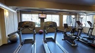 This fitness center offers a practical and well-appointed space for exercise and wellness. The single-floor layout is designed to maximize usability, providing ample room for a variety of workout routines. The cardio area is outfitted with Life Fitness treadmills and a stationary recumbent bike, all positioned to take advantage of the natural light streaming in through large windows that look out onto surrounding greenery — creating an open, refreshing atmosphere during workouts. A wall-mounted flat-screen television is centrally positioned above the equipment, offering entertainment while exercising. The flooring features durable, cushioned blue fitness mats, providing both comfort and safety underfoot. Additional equipment, including a medicine ball and strength training machines, rounds out the available amenities. The walls are clad in warm wood paneling, lending a classic, welcoming feel to the space. Overhead, the ceiling incorporates ventilation to maintain air circulation throughout workouts. The overall ambiance is functional yet comfortable, striking a balance between practicality and a motivating environment suited for both light and moderate fitness activities.
